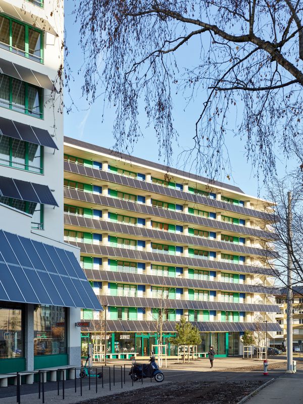 Areal Guggach – Die Platzfassade mit ausladenden PV-Modulen und kräftigen Natursteinstützen wird zu «Stadtloggia» mit Alpenpanorama. Auch im Erdgeschoss bilden Sitzbänke und Vordächer geschützte Begegnungs- und Pausenräume entlang der vielbefahrenen Strasse. – Philip Heckhausen